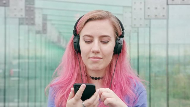A young woman with a pink hair wearing headphones using a phone in the city street. Close-up shot. Soft focus