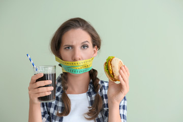 Emotional woman with measuring tapes around her mouth and unhealthy food on color background. Diet...