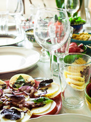 Served banquet table for festive dinner with empty plates, fork, knives and wine glasses. In the foreground Caprese salad: tomato, mozzarella, onion, basil and balsamic vinegar. Day light. Soft focus.