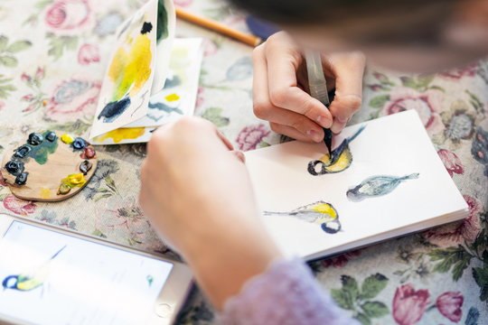Teenager girl drawing bird in small paper drawing-pad. Close-up kid artist painting small picture on paper notepad with brush. Children creativity concept
