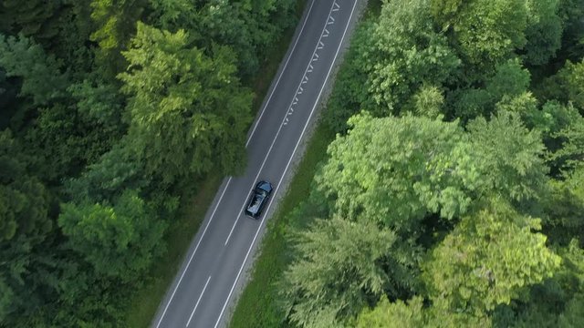 AERIAL, TOP DOWN: Car driving through the woods gets overtaken by a fast motorbike rider. Aerial shot of a dark car and bike driving down forest road on a sunny day. Relaxing cruise in the woods.