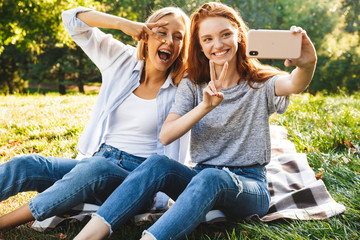 Image of two smiling girls wearing casual clothes sitting on grass and showing peace sign, while taking selfie on smartphone in summer park