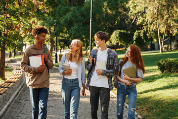 Multiethnic young students guys and girls smiling, while walking together in park with books