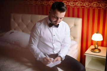 Handsome groom sitting on the bed in the morning in the room. Young man in white shirt with bow tie...