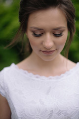 Naklejka premium Portrait of young beautiful bride woman with nude make up in white wedding dress lookimg down outdors in the park. Wedding concept. Close up