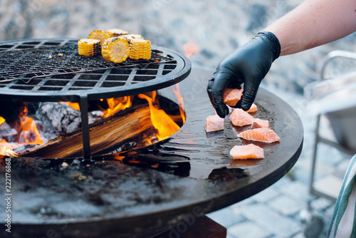 Man Cooking Corn And Salmon On The Big Round Flaming Bbq Grill Man Cooking Corn And Salmon On The Big Round Flaming Bbq Grill Outdoors Open Fire
