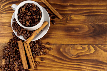 Coffee beans in white cup and cinnamon sticks on wooden table. Top view, copy space