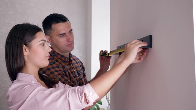 smiling married couple hanging shelf on wall with tape measure in arms during repairs in new accommodation