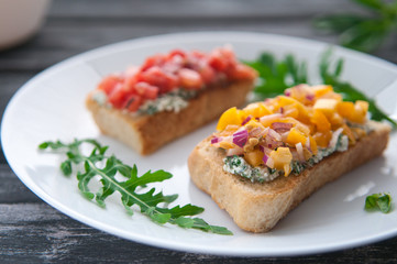 Bruschetta with cheese red and yellow tomatoes on a white plate. The composition is decorated with leaves of basil, arugula, pepper and vegetables. Side view on an old wooden background. Blurred.