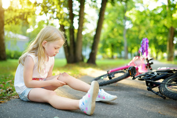 Cute little girl sitting on the ground after falling off her bike at summer park. Child getting...
