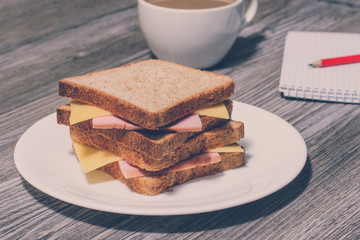 Business breakfast. Tasty ham and cheese sandwich with cup of coffee, notebook and pencil. Gray wooden background, vintage effect
