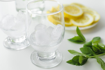 Two transparent glasses with ice cubes, fresh mint leaves on white table and lemon slices on a plate in the background.