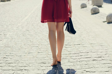Young pretty woman in red dress walking in the city without shoes, she is holding high-heels in her hand. Close up photo of woman's long barefoot legs against road, view from back