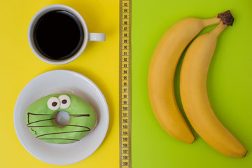 Concept of unhealthy eating and snack. Delicious donut and cup of strong coffee isolated on yellow and fresh tasty bananas isolated on green background. Top view photo