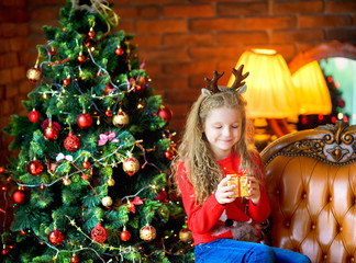 beautiful girl with a small gift sits in a chair near a festive Christmas tree.