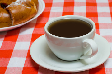 Cup of coffee on checkered red tablecloth with cake on the background. Close up photo