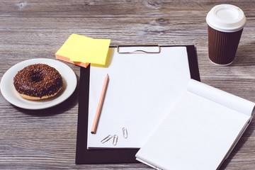Overhead view of office accessorise: clipboard, tasty donut, paper cup of coffee, notepad and clipboard