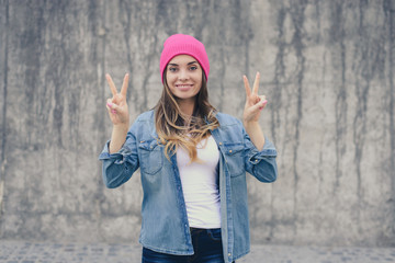 Happy joyful smiling hipster girl in casual clothes and pink hat standing against gray wall and...