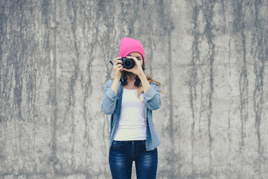 Hipster girl in jeans clothing and pink hat taking photo against grey concrete wall. Focus on camera