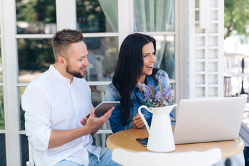 The concept of gadgets, technology, freelancing, people. A young European man sits at a table in a cafe with his girlfriend and talks about something, laughs. Friends rest in the restaurant outside