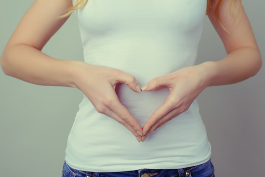 Concept of first weeks pregnancy. Woman's hands forming heart on her tummy. Concept of healthy eating and lifestyle. Woman in white t-shirt and jeans isolated against grey background