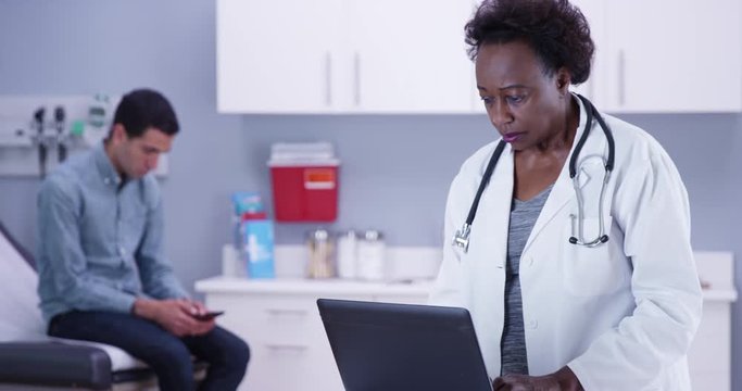 Close view of female senior African doctor typing notes on latop computer of male patient. Portrait of black MD using portable computer to record notes of young male patient
