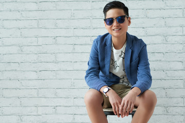 Cheerful Asian cool boy in sunglasses sitting on chair at brick wall and looking at camera