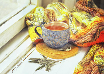 Cup of black tea, sprigs of sage and knitted blanket on windowsill.