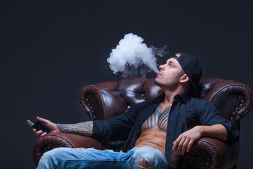 Vaper. The man with tattoos sits on a leather sofa smoke an electronic cigarette on the dark...