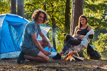 Young couple of hikers sitting warming near a campfire at camp in the forest on sunny day.