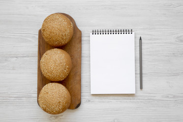 Fresh burger buns with sesame seeds on wooden board and blank notepad on white wooden background, overhead view. Flat lay, from above, top view.