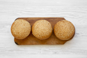 Fresh burger buns with sesame seeds on wooden board on white wooden background, top view. Fast food.