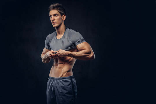 Stylish ectomorph bodybuilder with stylish hair takes off his shirt on the dark background.