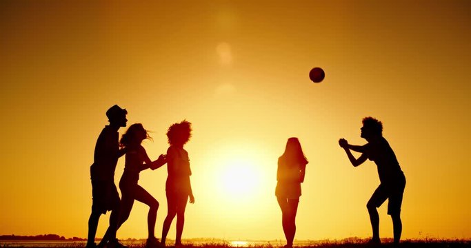 Silhouettes of active friends playing volleyball on a meadow near lake at sunset, and cheering. Friends having fun 4k