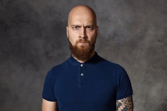 What's your problem? Picture of unfriendly enraged young bearded macho man with shaved head frowning and pursuing lips, being angry with something, his eyes full of rage, indignation and anger
