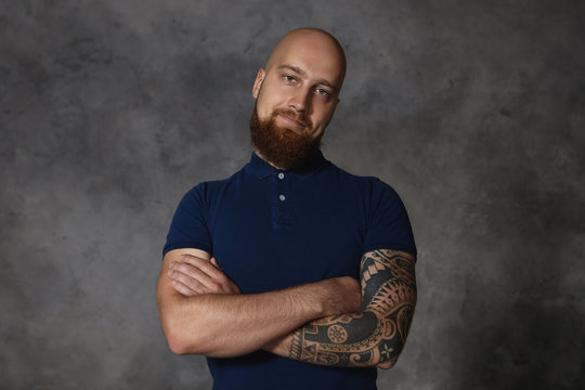 Horizontal isolated picture of stylish handsome young European guy with shaved head and fuzzy ginger beard keeping his arms folded and looking at camera with skeptical smile, bending head to one side