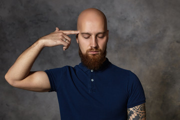 Boredom, fatigue and depression concept. Isolated shot of depressed unhappy Caucasian male with thick beard holding fore finger at his temple and closing eyes as if shooting, feeling stressed