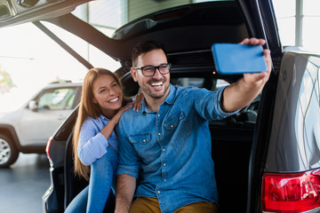 Beautiful excited young couple at car showroom taking selfie photo in their new car.