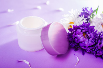 Fototapeta na wymiar Jar with cream surrounded with flowers on purple background. Organic cosmetics. Bodycare