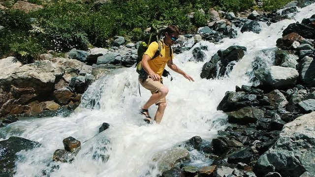 Hiker tourist climber man with backpack cross a cold mountain stream into a ford, slow motion. Extreme hiking