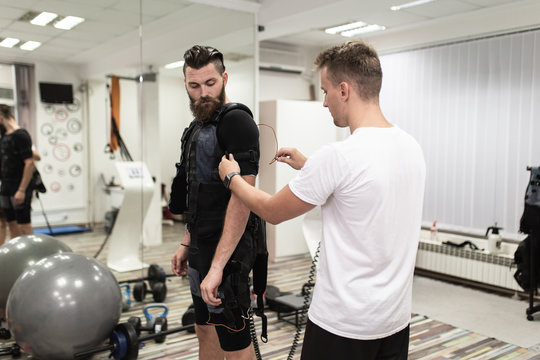 Young bearded man doing exercises in electrical muscular stimulation suit with his personal trainer at rehabilitation center.