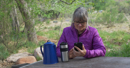 Elderly female hiker resting on bench reading smartphone outdoors in nature