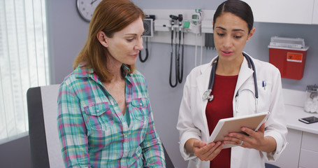 Fototapeta premium Charming female senior patient consulting with young latina doctor about health condition. Portrait of young hispanic doctor using portable tablet to look at test results of mid aged patient