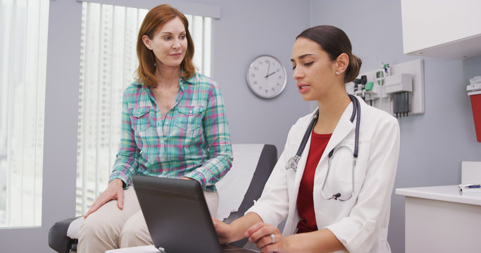 Portait Of Young Latina Doctor Using Laptop Computer While Talking With Senior Patient Inside Clinic. Close Up Of Middle Aged Patient Discussing With Young Female Doctor Health Issues