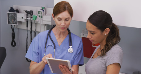 Portrait of middle aged female nurse using tablet computer to show young latina patient her medical...