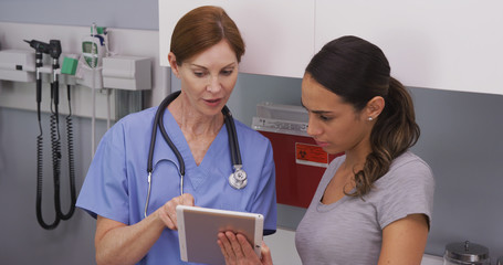 Portrait of middle aged female nurse using tablet computer to show young latina patient her medical...