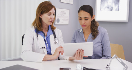 Senior medical physician using high-tech tablet to share lab results with young patient. Young patient consulting with mid aged doctor her health conditions looking at tablet computer