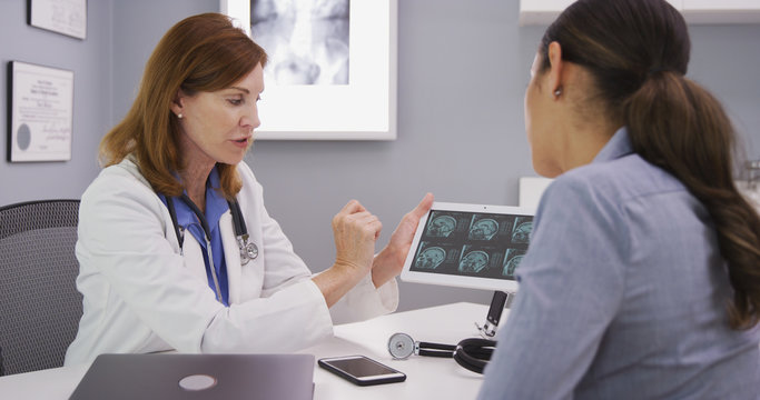 Young attracive latina patient reviewing ct scan of cranium with middle aged doctor.  Senior doctor consulting with patient over mri images on high tech tablet