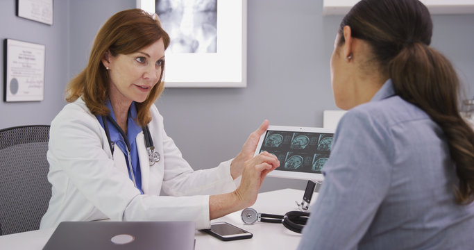 Young attracive latina patient reviewing ct scan of cranium with middle aged doctor.  Senior doctor consulting with patient over mri images on high tech tablet
