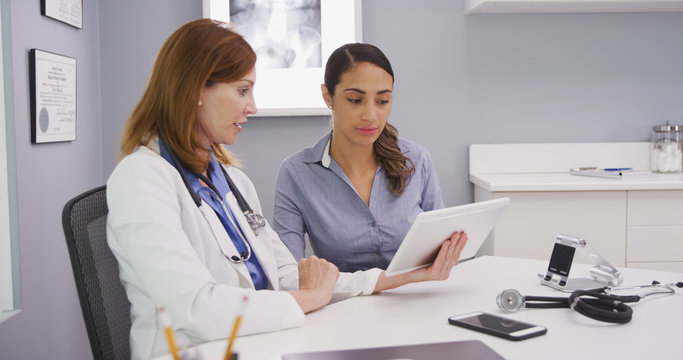 Charming Female Senior Doctor Using Tablet Computer To Share Lab Results With Young Latina Patient. Professional Medical Physician And Patient Looking At Tablet Portable Device